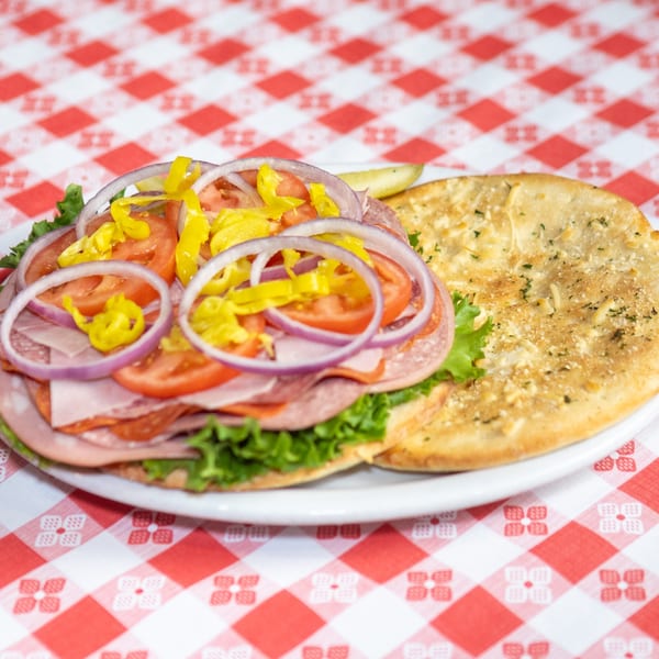 Open-faced Italian sub with salami, ham, lettuce, tomatoes, onions, and banana peppers on a red and white checkered tablecloth.