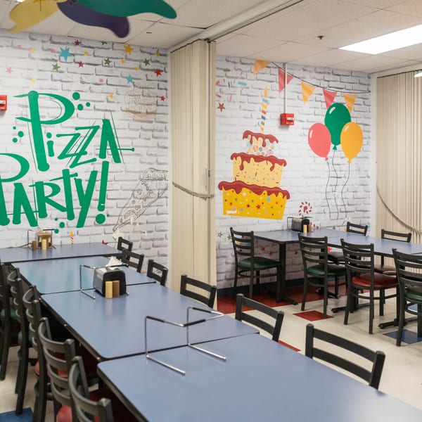 A colorful party room with blue tables, black chairs, and festive wall murals of pizza and cake.