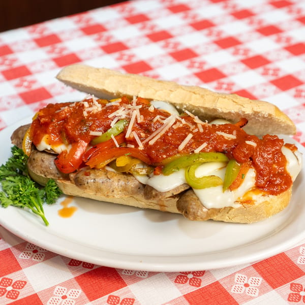 Sausage and pepper hero sandwich with melted cheese, marinara sauce, and parsley on a checkered tablecloth.