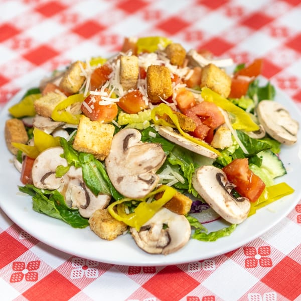 Fresh garden salad with mushrooms, tomatoes, croutons, and peppers on a white plate over a red and white checkered tablecloth.
