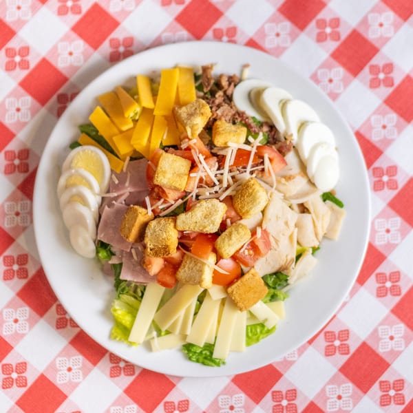 Chef salad with ham, turkey, cheese, tomatoes, croutons, and hard-boiled eggs on a red and white checkered tablecloth.