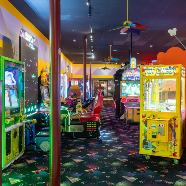 A brightly lit arcade room with various video games, claw machines, and a colorful patterned carpet.