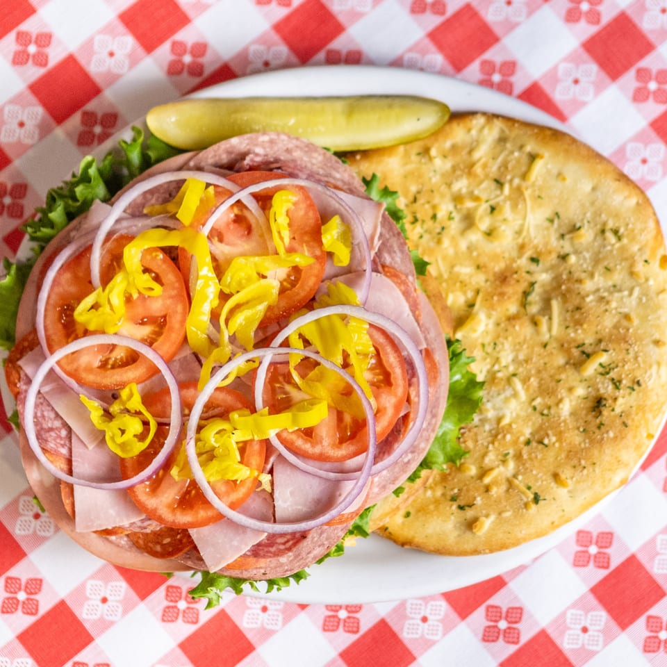 Italian sub sandwich with lettuce, tomato, onion, and peppers, served with a pickle and flatbread.
