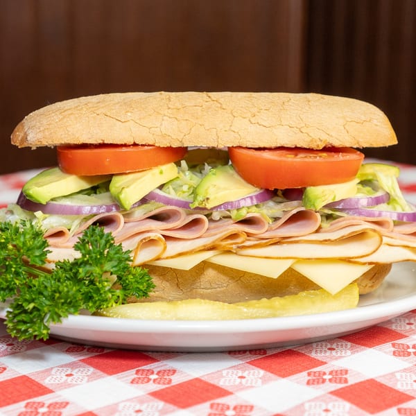 A large deli sandwich with turkey, cheese, avocado, tomato, and red onion on a white plate.