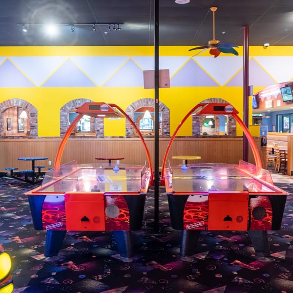 Two red air hockey tables with blue mallets in a brightly lit arcade room with yellow walls.
